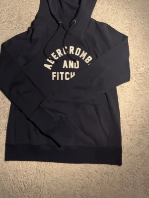 Blå hoodie från Abercrombie & Fitch - Mörkblå hoodie från Abercrombie & Fitch med vit text på bröstet! Riktigt snygg o skön knappt använd alls. Skriv vid minsta fundering!!!