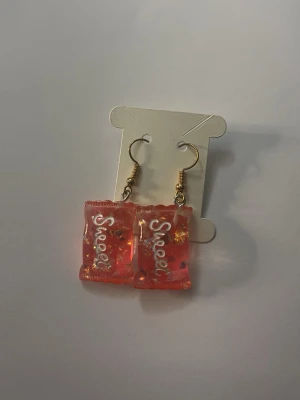 Rosa godisörhängen med text - Unika örhängen formade som små godispåsar i rosa plast med texten 'Sweet' på framsidan. Örhängena har guldfärgade krokar och transparent design med små dekorativa detaljer inuti. Perfekta för dig som gillar lekfulla accessoarer.