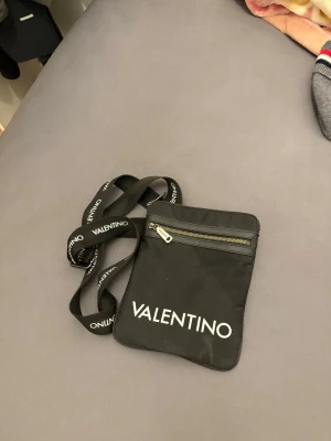 Svart axelväska från Valentino - Snygg svart axelväska från Valentino med justerbart axelband där märkesnamnet står tryckt i vitt. Väskan har en dragkedjeficka framtill och stor logga på framsidan. Perfekt för att bära det viktigaste och ger en trendig streetstyle-look.