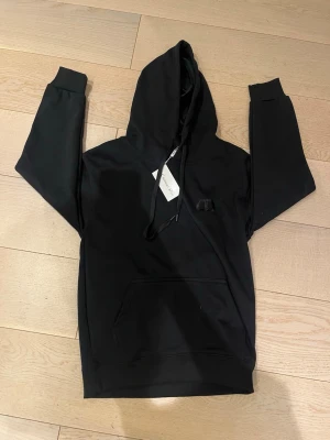 Svart märkeshoodie  - Snygg svart hoodie från CD med klassisk huva och dragsnören. Diskret broderad CD-logga på bröstet och stor magficka framtill. Tillverkad i mjukt bomullsmaterial som känns skönt mot huden. Perfekt för en avslappnad och stilren look.