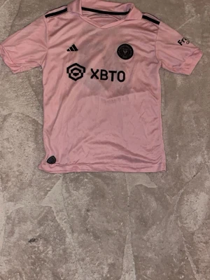 Rosa Inter Miami Messi matchtröja - Säljer en rosa Inter Miami matchtröja från Adidas med Messi och nummer 10 på ryggen. Tröjan har svarta detaljer, klubbmärke och sponsorlogga framtill samt korta ärmar. Perfekt för fotbollsfans som vill sticka ut på planen eller läktaren.