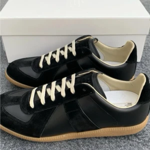 Svarta Maison Margiela Replica sneakers - Svarta Maison Margiela Replica sneakers med ovandel i läder och mocka, kontrasterande vita snören och beige innersula. Klassisk gummisula i ljusbrun färg och diskret branding på plösen. Snygg och tidlös modell med låg profil.