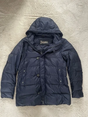 Mörkblå dunjacka från Herno med huva - Snygg mörkblå dunjacka från Herno med fast huva och knappar framtill. Jackan har två sidfickor med knappstängning och quiltad design. Perfekt för kalla dagar när du vill hålla dig varm och stilren. Klassisk passform och lätt glansigt material.