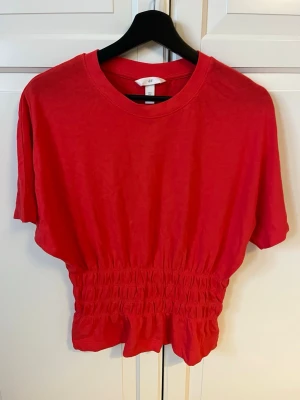 Röd croppad topp från H&M - Säljer en röd croppad topp från H&M med korta ärmar och rund hals. Toppen har smockad midja som ger en snygg passform och är tillverkad i mjuk bomull. Perfekt att styla med höga jeans eller kjol.