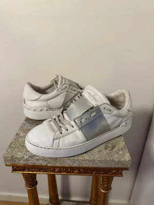 Valentino Open sneakers vit/silver - Snygga Valentino Open sneakers i vitt skinn med en bred metallic-silver detalj över mitten. Klassisk rund tå, platt sula och snörning. Ikoniska nitar baktill ger extra edge. Perfekta för dig som gillar stilrena men ändå unika sneakers.