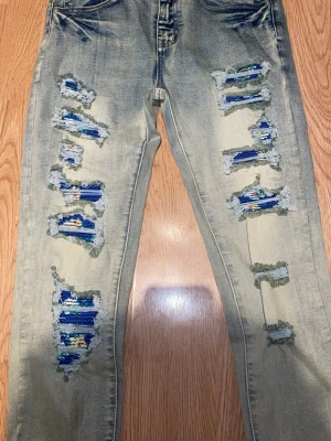 Ljusblå/grå slitna jeans med färgglada lappar - Unika ljusblå jeans med kraftiga slitningar och färgglada lappar i blått, grönt och gult under de trasiga partierna. Jeansen har klassisk femficksmodell och raka ben. Perfekt för dig som gillar en edgy och personlig stil.