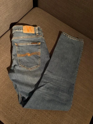 Blå jeans från Nudie Jeans - Säljer ett par klassiska blå jeans från Nudie Jeans slim fit och snygga slitningar framtill. Jeansen har orange sömmar och en ikonisk bakficka med vågig brodyr. Tillverkade i mjuk denim, perfekta för en avslappnad stil.