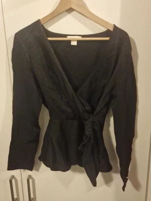 Svart omlottopp med knytband - Snygg svart omlottopp med knytband i midjan och v-ringad hals. Toppen har långa ärmar och lätt glittrig yta som ger en extra touch. Figurnära passform och lätt utsvängd nedtill. Perfekt för dig som vill ha något stilrent men ändå speciellt.