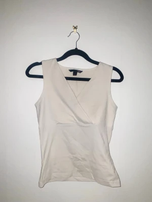 Vit omlottopp - Snygg vit omlottopp med v-ringning och ärmlös design. Toppen har en figurnära passform, perfekt för en clean och stilren look. Passar till både jeans och kjol för en fräsch outfit.