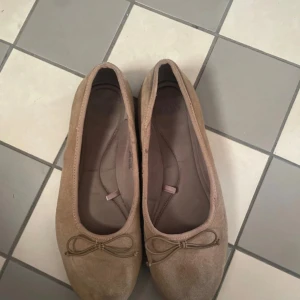 Beige/brun ballerinaskor med rosett - Säljer ett par klassiska beige ballerinaskor i mocka med rund tå och söt rosettdetalj framtill. Skorna har en enkel och stilren design som passar till många olika outfits. Perfekta för dig som gillar en clean och tidlös look.