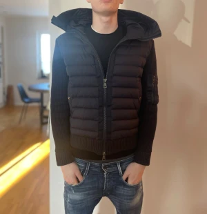 Svart dunväst från Moncler - Snygg svart dunväst från Moncler med stickade ärmar och huva. Västen har dragkedja framtill, ribbade muddar och en ficka med Moncler-logga på ärmen. Perfekt för lager-på-lager och ger en sportig look.