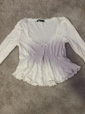 Vit långärmad blus med spetsdetaljer - Söt vit blus med lång ärm, spetskant längs ringningen och framtill samt liten rosett vid bysten. Modellen har en lätt utsvängd midja och volangkant nedtill. Perfekt för en romantisk och feminin stil.