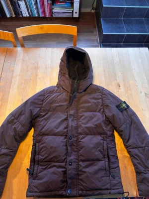 Brun dunjacka från Stone Island - Säljer en brun dunjacka från Stone Island med avtagbar patch på ärmen. Jackan har huva, dragkedja och knappar framtill samt två sidofickor. Perfekt för kalla vinterdagar och riktigt snygg streetstil. 100% äkta, kom privat för certilogo. Priset är förhandlingsbart 