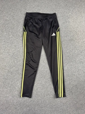 Svarta Adidas träningsbyxor med gula ränder - Säljer ett par svarta träningsbyxor från Adidas med klassiska gula ränder längs benen och vit Adidas-logga på låret. Byxorna har smal passform, resår i midjan och dragkedjor vid bensluten. Perfekta för träning eller chill.