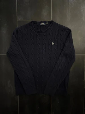 Mörkblå kabelstickad tröja Polo Ralph Lauren - Kabelstickad mörkblå tröja från Polo Ralph Lauren med klassisk rund halsringning och broderad logga på bröstet. Tröjan har långa ärmar och ribbade muddar vid ärmslut och nederkant. Perfekt för dig som gillar stilrena och tidlösa plagg.