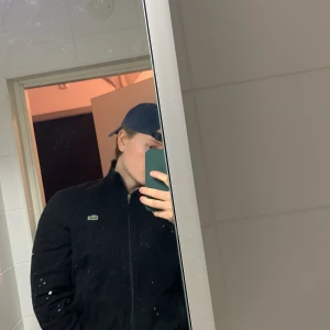 Svart ziptröja från Lacoste - Säljer en svart ziptröja från Lacoste med klassisk krokodillogga på bröstet. Tröjan har hög krage, dragkedja framtill och ribbade muddar. Perfekt för en stilren och sportig look. 