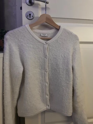 Vit fluffig kofta från Kappahl  - Mysig vit kofta från Kappahl med fluffig textur och pärlknappar framtill. Rund halsringning och ribbade kanter ger en clean look. Perfekt att slänga över en topp för extra värme och stil.