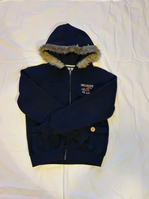 Marinblå hoodiejacka från Hollister - Mysig marinblå hoodiejacka från Hollister med fuskpäls runt huvan och dragkedja framtill. Jackan har broderad logga på bröstet, två fickor med knappdetalj och ribbade muddar. Perfekt för kyliga dagar.