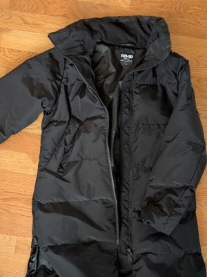 Svart lång jacka 8848 Altitude - Snygg svart lång jacka från 8848 Altitude med hög krage och dragkedja framtill. Jackan har en stilren design och är perfekt för kalla vinterdagar. Tillverkad i slitstarkt material med värmandefyllning och diskret logga på bröstet. Ålder 10-12 år.