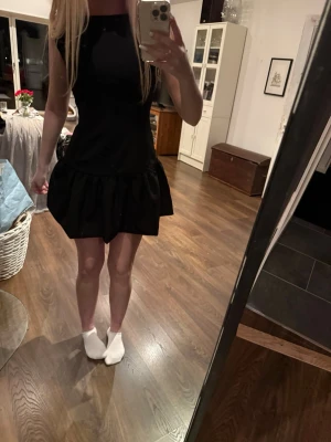 Svart kort klänning med volang - Snygg svart kort klänning med volang nertill och ärmlös design. Klänningen har en enkel, stilren form och passar perfekt för dig som gillar en klassisk look. Materialet känns mjukt och följsamt mot kroppen.