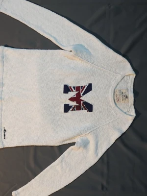 Vit tröja med M och Union Jack - Vit långärmad tröja från Morris med ett stort M på bröstet i Union Jack-mönster. Tröjan har rund hals och är tillverkad i ett mjukt, stickat material. Perfekt för en avslappnad och stilren look.