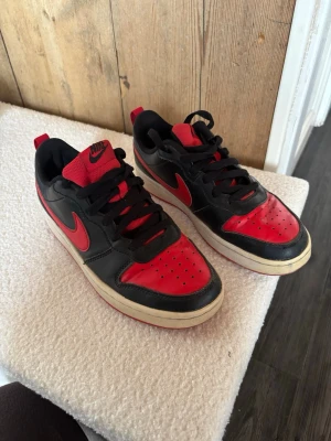 Nike svart-röda sneakers låga - Snygga Nike sneakers i svart och rött med klassisk swoosh på sidan. Skorna har lågt skaft, svarta snören och röd plös med Nike-logga. Ovandelen är i läder och textil, med perforerade detaljer framtill för extra ventilation.