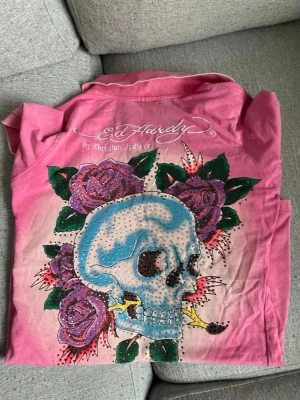 Rosa Ed Hardy skjorta med dödskalle - Säljer en rosa Ed Hardy skjorta med ikoniskt dödskalle- och ros-tryck både fram och bak. Skjortan har krage, knappar framtill och coola stenar som ger extra bling. Materialet är mjuk bomull och designen är riktigt statement med färgstarka detaljer.