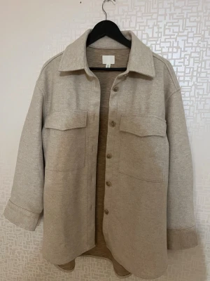 Beige overshirt från H&M - Stilren beige overshirt från H&M med klassisk krage, stora bröstfickor och knäppning framtill. Overshirten har en rak passform, Storlek M men passar även L, aldrig använd bara testad