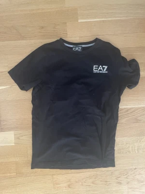 Svart EA7 t-shirt med tryck - Snygg svart t-shirt från EA7 Emporio Armani med vitt logotryck på bröstet. Klassisk rund hals och korta ärmar. Perfekt för dig som gillar stilrena och enkla plagg med en exklusiv touch.