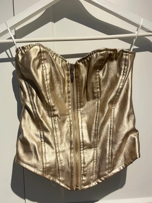 Guldig bustierkorsett med dragkedja - Snygg guldig korsett i glansigt material med dragkedja framtill och markerade sömmar för en strukturerad look. Axelbandslös design och lätt vadderad för extra stöd. Perfekt för att lyfta vilken outfit som helst.