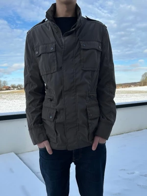 Mörkgrön fieldjacket med fickor - Snygg mörkgrön fieldjacket med fyra stora fickor framtill och hög krage. Jackan har dragkedja och tryckknappar, samt axelklaffar för extra stil. Perfekt för kyligare dagar och har en klassisk, rak passform.