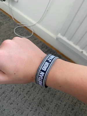 Christian Dior J'adior armband - Snyggt vävt armband från Christian Dior i marinblått och vitt med texten 'CHRISTIAN DIOR J'ADIOR'. Armbandet har justerbar knytning och dekorativa fransar i ändarna. Perfekt accessoar för att lyfta din outfit.