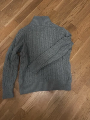 Grå kabelstickad tröja Polo Ralph Lauren - Snygg grå kabelstickad tröja från Polo Ralph Lauren. Tröjan har hög krage, långa ärmar och ribbade muddar vid ärmslut och nederkant. Perfekt för dig som gillar klassisk och stilren design.