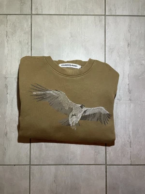 J.lindberg tröja  - Beige tröja från J.Lindeberg Denim med ett stort stilrent tryck på bröstet. Tröjan har rund halsringning och ribbade muddar. Perfekt för dig som gillar en tillbaka lutad stil. Passar strl: M-L