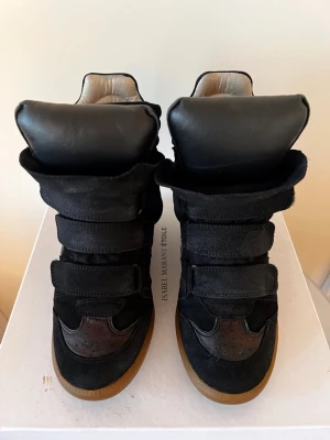 Svarta wedge sneakers Isabel Marant - Snygga svarta wedge sneakers från Isabel Marant med högt skaft, kardborreband och gummisula. Skorna är gjorda i mocka och läder med coola detaljer och en chunky siluett. Perfekta för dig som vill ha en edgy och trendig look.