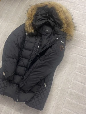 Svart dunjacka med fuskpäls från Rockandblue - Snygg svart dunjacka från Rockandblue med quiltade detaljer och avtagbar huva med fuskpäls. Jackan har dragkedjor på fickorna, emblem på ärmen och en figurnära passform. Perfekt för kalla vinterdagar.