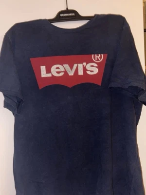 Blå Levi's t-shirt med logga - Säljer en mörkblå t-shirt från Levi's med den klassiska röda loggan tryckt på bröstet. T-shirten har korta ärmar och är gjord i mjuk bomull, perfekt för en avslappnad och stilren look. Ikonisk röd Levi's-tag på sidan.