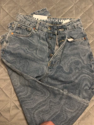 Blå mönstrade wide leg jeans från Agolde - Säljer ett par ljusblå wide leg jeans från Never denim med unikt vågigt mönster i tyget. Jeansen har hög midja, knäppning med knappar och klassiska fem fickor. Perfekta för dig som vill sticka ut med en cool och trendig look.