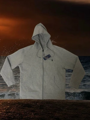 Grå hoodie från Polo Ralph Lauren - Snygg grå hoodie från Polo Ralph Lauren med dragkedja framtill och klassisk huva med dragsko. Mjuk bomullsblandning, två fickor fram och diskret broderad logga på bröstet. Perfekt för en chill och stilren look.