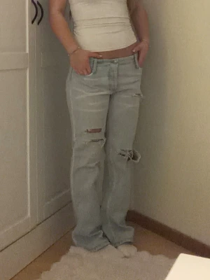 Ljusblå slitna raka jeans - Säljer ett par ljusblå jeans med raka ben och flera slitningar på framsidan. Jeansen har hög midja och klassisk femficksdesign. Perfekta för en avslappnad och trendig look.