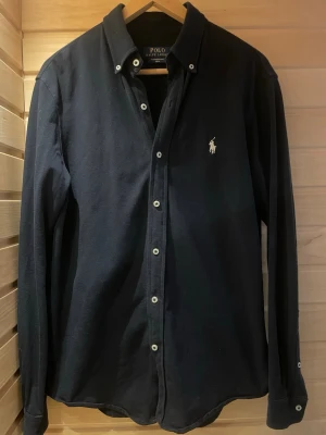 Svart skjorta från Polo Ralph Lauren - Klassisk svart skjorta från Polo Ralph Lauren med vit broderad logga på bröstet. Skjortan har lång ärm, knappar framtill och button-down krage. Tillverkad i mjuk bomull för en bekväm känsla.