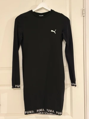 Svart långärmad klänning från Puma - Svart långärmad klänning från Puma med vit logga på bröstet och PUMA-tryck längs nederkant och ärmslut. Klänningen har en rak passform och är gjord i mjukt bomullsmaterial. Perfekt för en sportig och avslappnad stil.