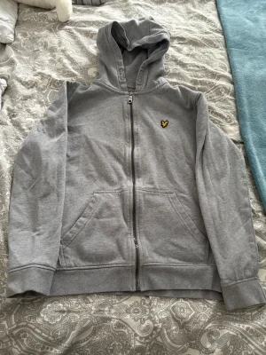 Grå zip hoodie från Lyle & Scott - Snygg grå hoodie med dragkedja från Lyle & Scott. Klassisk design med huva, två fickor framtill och den ikoniska gula örnloggan på bröstet. Tillverkad i mjukt sweatshirtmaterial som är skönt att ha på sig. Perfekt för en avslappnad stil.