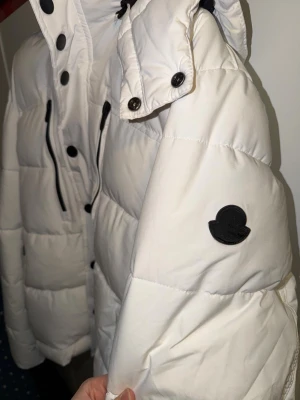 Vit dunjacka från Moncler - Snygg vit dunjacka från Moncler med svart logotyp på ärmen, svarta detaljer och dragkedjor. Jackan har hög krage, avtagbar huva och flera fickor med knappar och dragkedjor. Perfekt för kalla vinterdagar och riktigt varm.