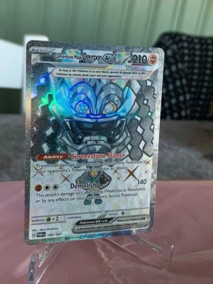 Ogerpon ex Pokémonkort holo - Säljer ett Ogerpon ex Pokémonkort med holografisk yta och färgstarka detaljer. Kortet har 210 HP och visar Ogerpon med ett maskliknande ansikte i blått, grått och svart. Kortet har specialförmågor och är från Scarlet & Violet-serien.