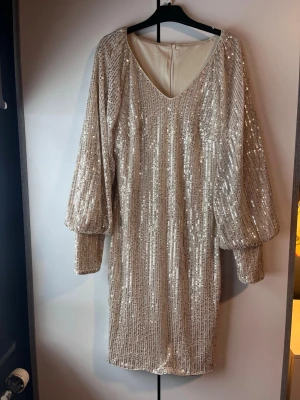 Glittrig kortklänning med paljetter - Säljer en glittrig kort klänning i champagnefärg med paljetter över hela klänningen. Klänningen har v-ringning och långa, puffiga ärmar. Perfekt för dig som vill sticka ut på festen!
