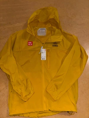 Gul vindjacka från Uniqlo SWE - Gul vindjacka från Uniqlo med huva och dragkedja. Jackan har SWE och OS-logga på bröstet samt Uniqlo-logga. Lätt material i polyester, perfekt för blåsiga dagar. Praktisk bröstficka med dragkedja och elastiska muddar vid ärmslut. Vindjackan är från OS i Paris
