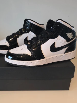 Air Jordan 1 Mid carbon svart/vit - Snygga Air Jordan 1 Mid carbon sneakers i svart och vitt med glansiga detaljer i lackläder och klassisk Swoosh på sidan. Skorna har en ikonisk siluett, vita paneler i textil och svarta partier i blankt material. Perfekta för dig som gillar streetwear och vill sticka ut. Nya i orginalkartong.