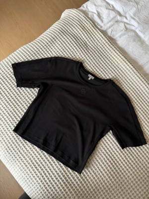 Svart croppad t-shirt från Loewe - Snygg svart croppad t-shirt från Loewe med diskret broderad logga framtill. T-shirten har en tightare modell, korta ärmar och är gjord i mjuk bomull, perfekt för en trendig och avslappnad stil. Använda fåtal gånger så i nyskick. 