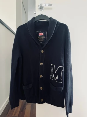 Svart kofta från Morris med M-logga - Snygg marinblå kofta från Morris med stora M-loggan på bröstet och dekorativa knappar framtill. Koftan har två fickor, sjalkrage och ribbade muddar. Perfekt för dig som gillar klassisk college-stil och vill ha något bekvämt och stilrent.